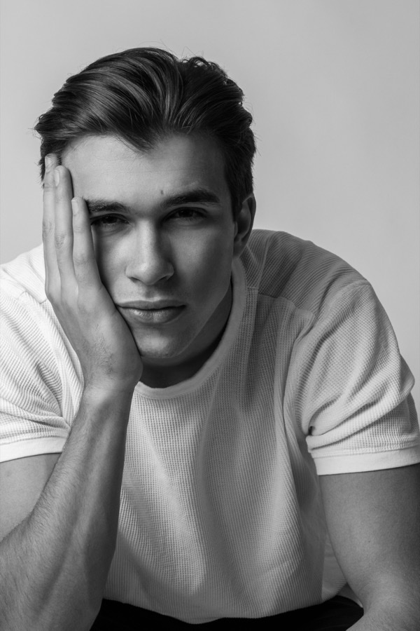 A black-and-white studio portrait of a young man resting his head on his hand