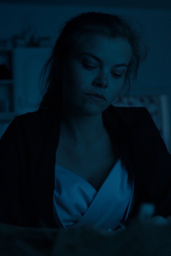 Film still — a woman sitting at a kitchen table at night, lit in cold blue light
