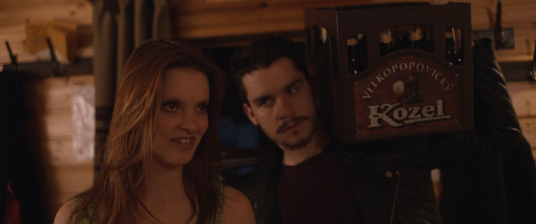 Film still — two characters in a warm, wood-panelled room with a crate of Kozel beer