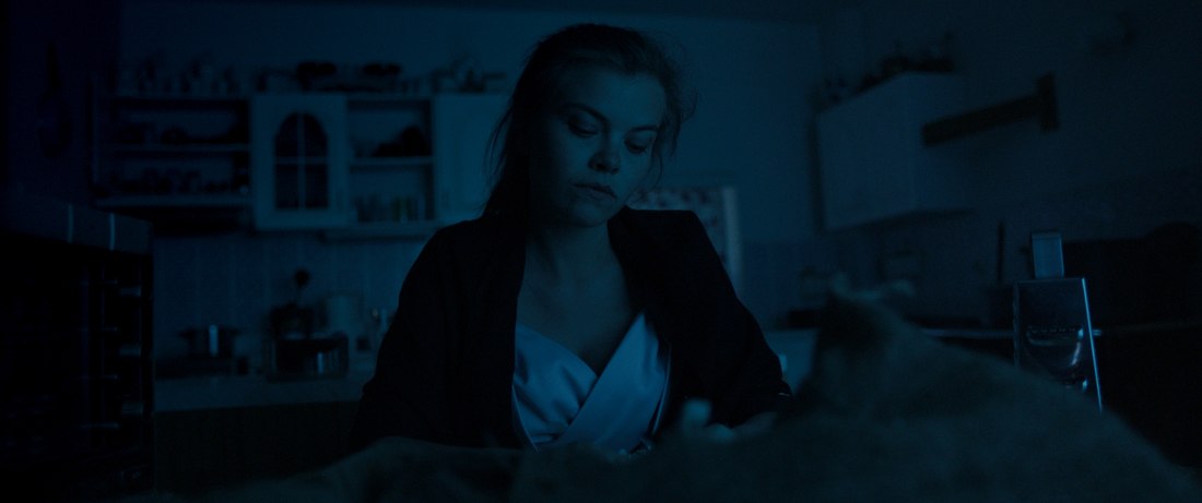 Film still — a woman sitting at a kitchen table at night, lit in cold blue light