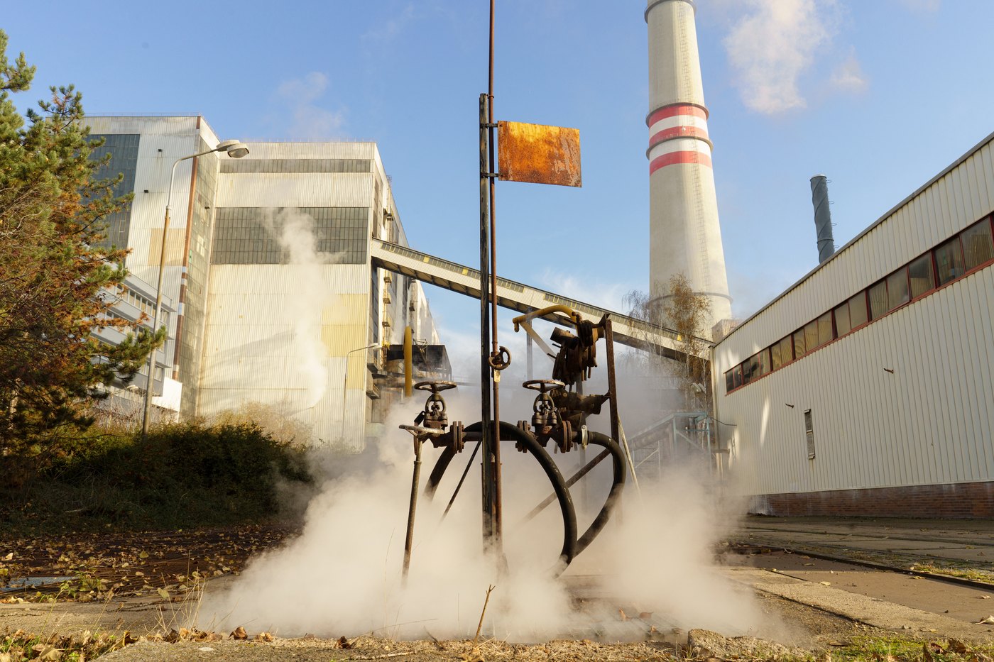 Steam venting from industrial valves and pipework, framed by the plant buildings and a smoking chimney behind