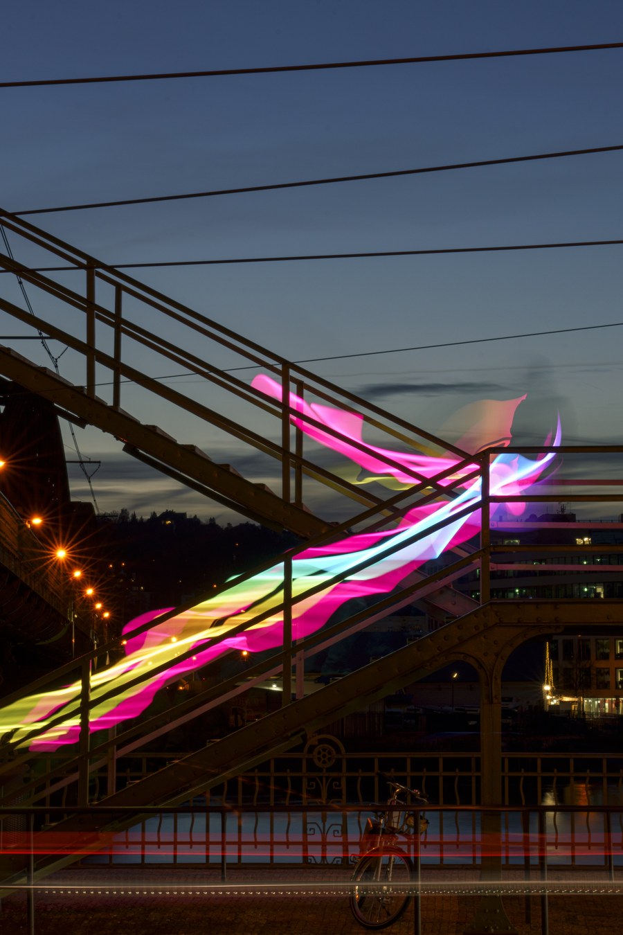 A long-exposure light-painting photograph — glowing pink, cyan and yellow ribbons of light sweeping across a steel bridge staircase at dusk
