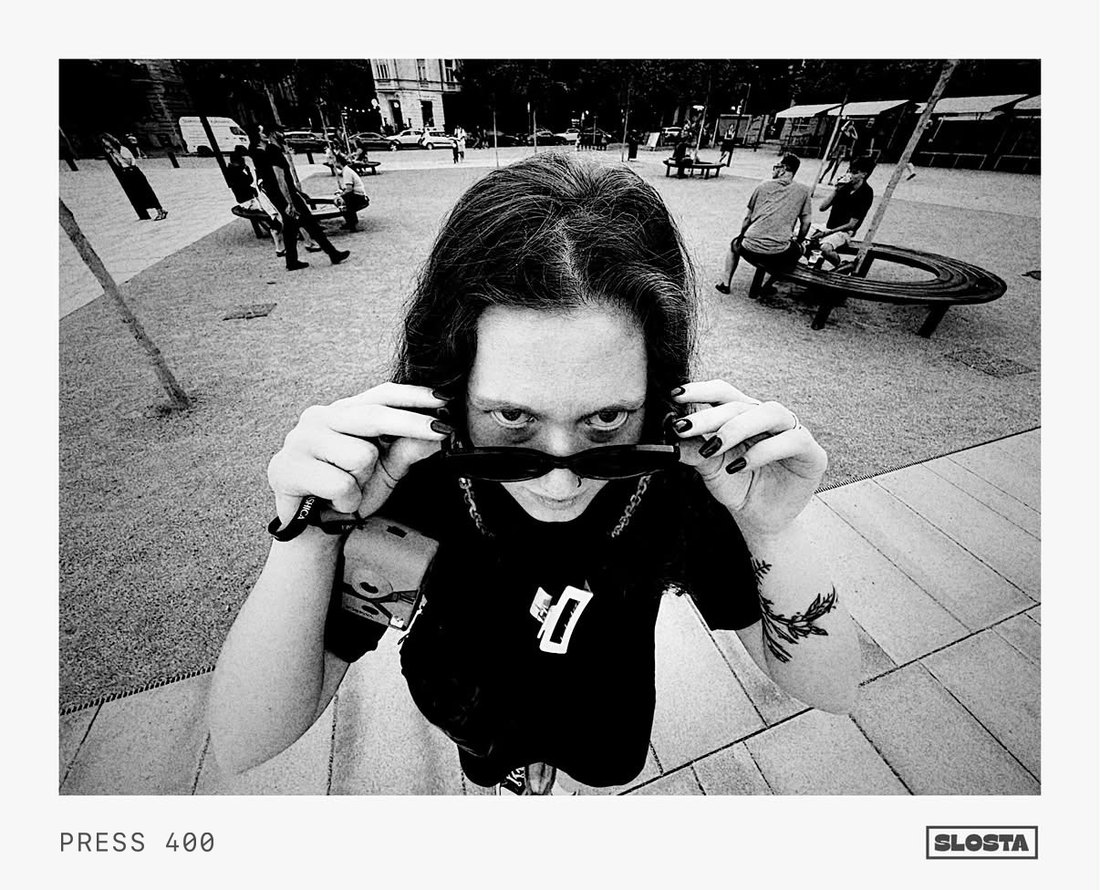 A person lowering their sunglasses to look at the camera in a city plaza, black-and-white film