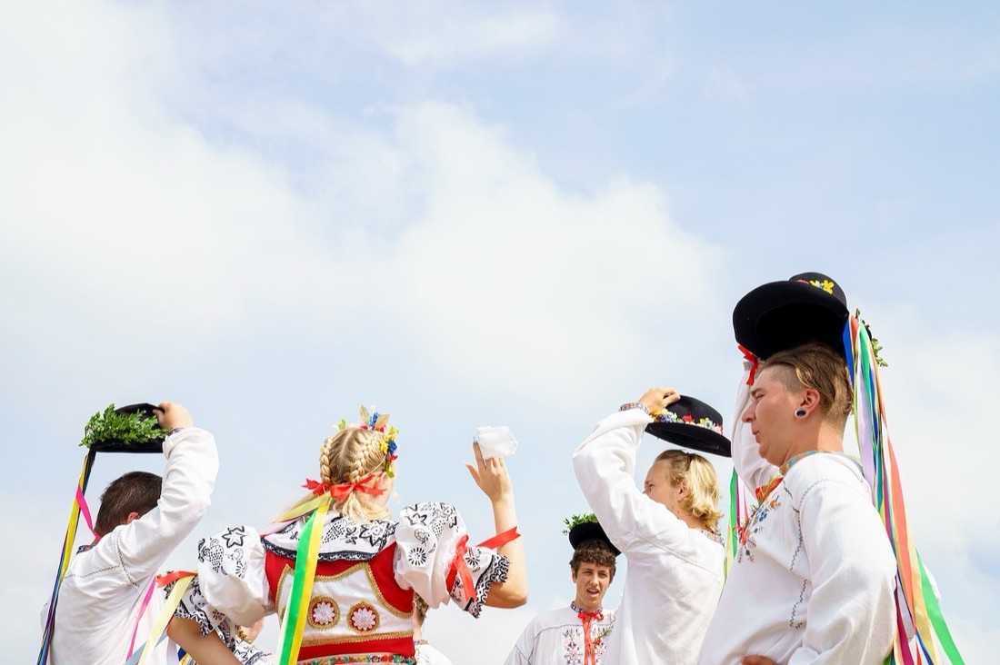 Young Moravian folk dancers in white embroidered shirts raising ribboned hats against a bright blue sky
